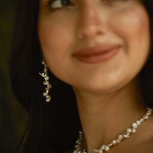 A woman with dark hair and a radiant smile wears an elegant keshi pearl jewelry set, featuring a delicate sterling silver pearl necklace and matching long dangle earrings.