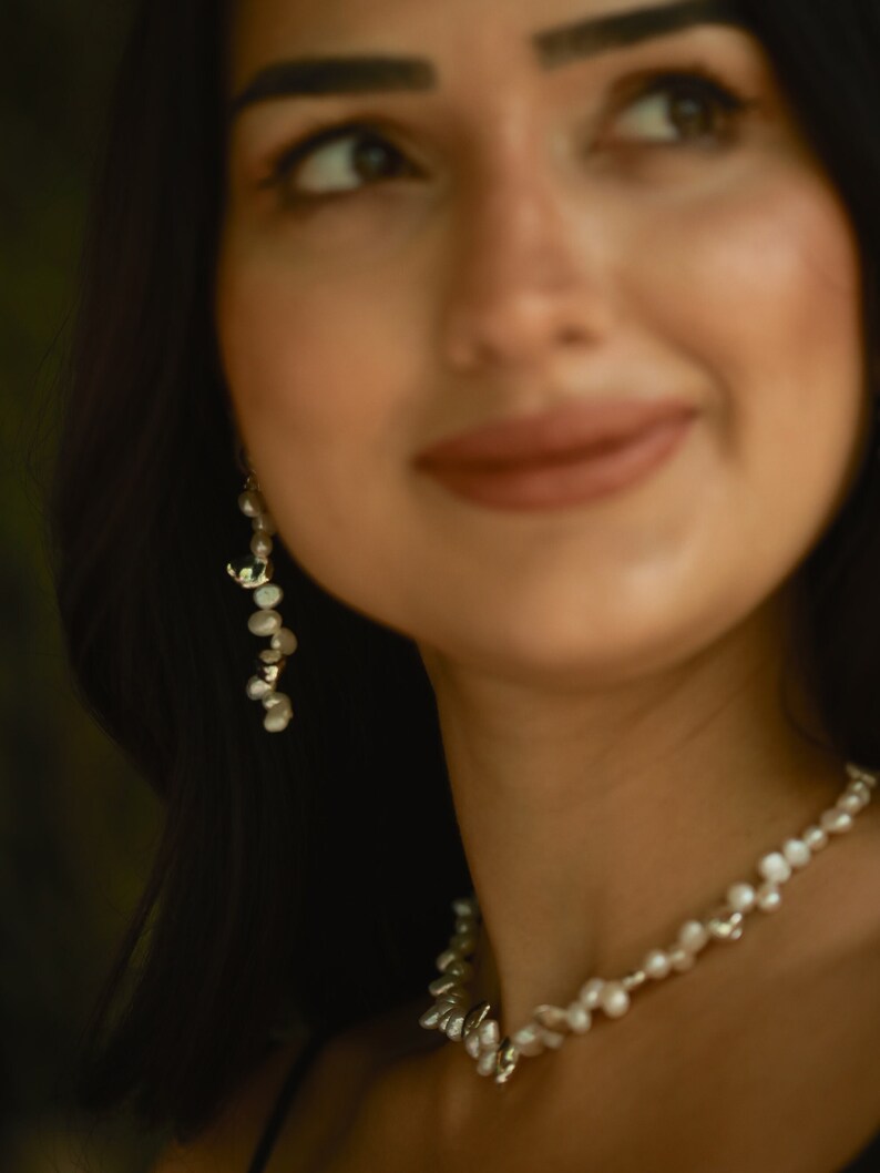 A woman with dark hair and a radiant smile wears an elegant keshi pearl jewelry set, featuring a delicate sterling silver pearl necklace and matching long dangle earrings.