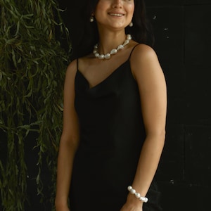 A woman is smiling while wearing a stunning baroque pearl jewelry set. The chunky necklace, matching bracelet, and drop earrings add a bold, elegant touch to her black dress. She stands in front of a leafy backdrop, giving the whole look a relaxed, natural vibe.