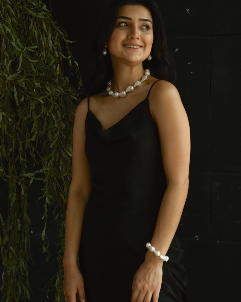 A woman is smiling while wearing a stunning baroque pearl jewelry set. The chunky necklace, matching bracelet, and drop earrings add a bold, elegant touch to her black dress. She stands in front of a leafy backdrop, giving the whole look a relaxed, natural vibe.