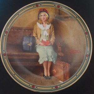 May include: A vintage-style porcelain plate with a gold rim and a green and gold border. The centre of the plate features a painting of a young woman in a brown coat and a green dress, sitting on a bench with a suitcase and a hatbox. The woman is holding a red box in her lap. The artist's signature, Norman Rockwell, is visible in the bottom right corner of the plate.