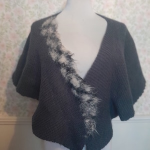 May include: A gray knitted shawl with a white and black fur trim. The shawl has a V-neck and short sleeves.