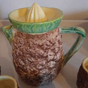 May include: A brown and green ceramic pitcher with a textured surface resembling a pineapple. The pitcher has a yellow citrus juicer on top and a green handle.