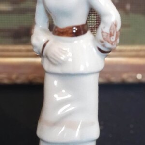 May include: A white porcelain figurine of a woman wearing a white dress and hat with brown accents. The figurine has a floral design on the skirt and is standing on a white base.