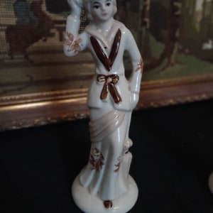May include: A white porcelain figurine of a woman wearing a sailor-style dress with brown accents. She is holding a small object in her hand and has a brown bow around her waist.
