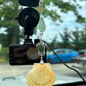 Könnte beinhalten: Ein hängendes Auto-Ornament mit einer hellgelben Jakobsmuschel an der Unterseite, einem silberfarbenen Meeresschildkröten-Anhänger mit grüner Emaille, einem Stück mattem hellgrünem Meerglas, einem kleinen goldfarbenen Seestern und weißen Perlen.