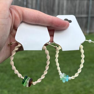 May include: Two macrame bracelets on white display cards. The left bracelet features twisted beige cord with dark green, light green, and brown sea glass beads. The right bracelet features twisted beige cord with light blue and green sea glass beads.