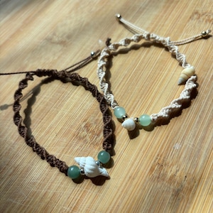 May include: Two adjustable macrame bracelets featuring twisted cord designs. One bracelet is dark brown with two green beads and a white seashell. The other is cream-coloured with two green beads, a small metallic bead, and a tiny seashell accent.