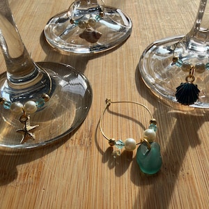 May include: Three wine glass charms featuring gold-tone wire hoops, white faux pearls, small light blue beads, and nautical-themed pendants including a starfish, a seashell, and a dark blue scallop shell, plus one loose sea glass charm.