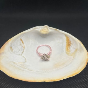 May include: A delicate ring made with pink wire and a small seashell charm. The ring is displayed on a white seashell.