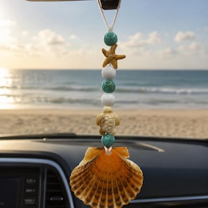 Könnte beinhalten: Ein hängendes Auto-Ornament mit einer vertikalen Schnur aus Perlen, einem kleinen Seestern, einem Meeresschildkröten-Anhänger und einer großen, fächerförmigen orange-braunen Muschel am unteren Ende. Die Perlen wechseln zwischen Türkis und Weiß.