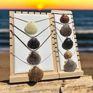 Puede incluir: Exhibición de collares de conchas marinas en soportes de madera. Los collares presentan diversas conchas en colores como blanco, negro, marrón y beige. Los soportes están colocados frente a una escena de playa con una puesta de sol al fondo.