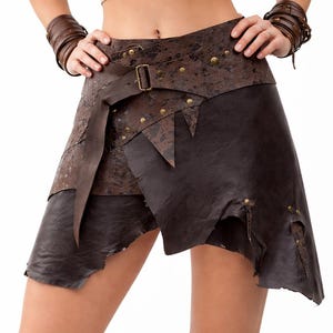 May include: A brown leather skirt with an unusual, asymmetrical design. The skirt has a belt with a buckle and is decorated with small, gold-coloured studs. The edges of the skirt have a distressed, rugged look, suggesting a rustic or fantasy theme.