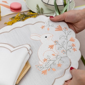 May include: White linen placemat with a scalloped edge featuring an embroidered white bunny rabbit sitting in a bush of pink and green flowers.