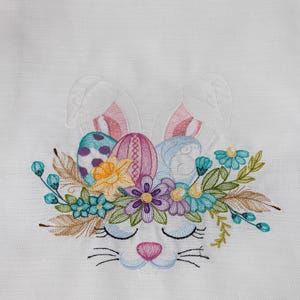 Op de afbeelding: Witte geborduurde keukendoek met een konijnenontwerp. Het konijn draagt een kroon van bloemen en paaseieren. Het ontwerp is in tinten roze, blauw, geel en groen.