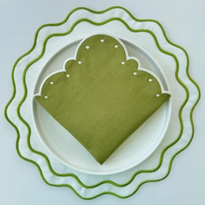 May include: A folded green napkin with white dotted trim rests on a white plate. The plate sits on a white placemat with a green wavy border. The placemat and napkin are part of a dining set.