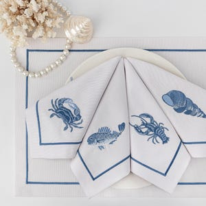 以下が含まれることがあります： カニ、魚、ロブスター、貝殻など、青い刺繍が施された白いリネンナプキン4枚。ナプキンは折り畳まれ、白いプレートの上に置かれています。