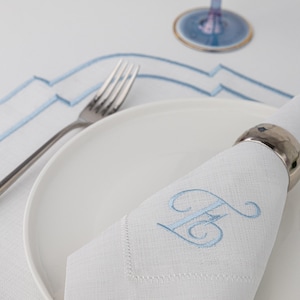May include: A white linen napkin with a blue embroidered letter "H" on it. The napkin is folded and placed on a white plate with a silver fork beside it.