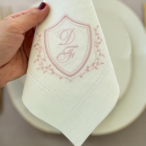 Puede incluir: Servilleta de lino blanco con un escudo bordado en rosa con las letras "D" y "F". El escudo está rodeado por un diseño floral. La servilleta está doblada y colocada sobre un plato blanco.