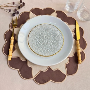 Könnte beinhalten: Tischarrangement mit einem weißen Teller mit Goldrand, auf einem strukturierten Glas-Unterteller. Goldene Gabel und Messer mit Bambusgriffen liegen auf einem braun-beigen Platzset. Zwei Gläser sind ebenfalls zu sehen.