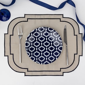Peut inclure: Un set de table beige avec une bordure brodée noire et un motif géométrique. Une assiette blanche avec un motif ikat bleu et blanc est posée sur le set de table, ainsi qu'une fourchette et un couteau.