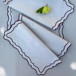 Puede incluir: Dos servilletas de lino blanco con bordes festoneados y un borde bordado de color morado oscuro. Las servilletas están dobladas y superpuestas.