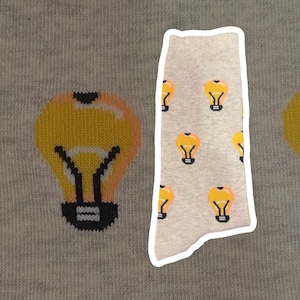 Könnte beinhalten: Hellgraue Socken mit einem Muster aus gelben Glühbirnen. Die Glühbirnen haben orangefarbene und schwarze Details. Die Socken werden auf einem hellgrauen Hintergrund präsentiert. Die Socken sind aus einem weichen, bequemen Material.