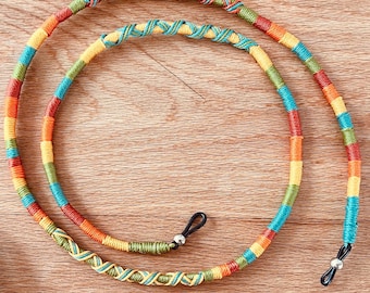 May include: A colorful eyeglass chain featuring a braided cord design in vibrant shades of orange, yellow, green, and turquoise. The cord includes small silver-toned metal beads and black rubber loops at each end for attachment to eyewear frames.