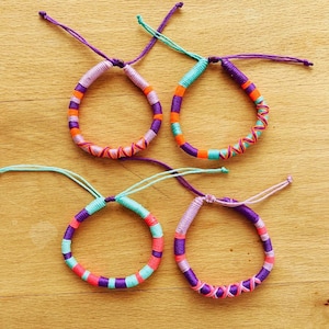 Può includere: Quattro braccialetti dell'amicizia fatti a mano con filo colorato. Ogni braccialetto presenta una combinazione unica di fili viola, arancioni, rosa e turchese, creando un design vivace. I braccialetti sono legati con cordini regolabili.