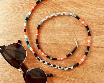 May include: Black sunglasses with gold accents rest on a wooden surface next to a colorful eyeglass chain. The chain features orange, black, white, and turquoise woven sections, and a silver loop for attachment.