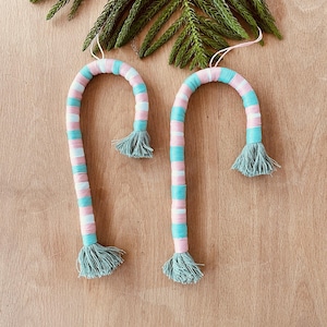 May include: Two candy cane-shaped ornaments wrapped in alternating stripes of light pink, white, and turquoise thread. Each ornament features a small, sage green tassel at the bottom end and a thin string loop at the top for hanging purposes.