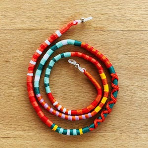 Peut inclure: Cordon de lunettes coloré, rouge, orange, jaune, vert et blanc. Le cordon est enroulé et possède des boucles transparentes pour l'attache aux lunettes. Le support est enroulé sur une surface en bois. Environ 70 cm de long.