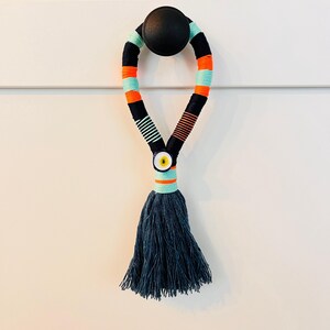 Può includere: Un tirare a frangia colorato con un design a strisce nero, arancione e turchese. La frangia è attaccata a un pomo nero e presenta un ciondolo blu a forma di occhio turco.
