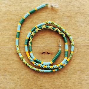 May include: A colorful eyeglass chain with a woven design. The chain features alternating sections of light blue, green, and yellow thread, with orange accents. The ends have clear loops for attaching to glasses.