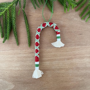 May include: A handmade Christmas ornament shaped like a candy cane. The ornament is red with a green and white crisscross pattern and white tassels at each end. A green string is attached for hanging. The ornament is displayed on a wooden surface with green pine needles.
