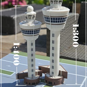 Könnte beinhalten: Zwei weiße und braune Modell-Flughafen-Kontrolltürme, einer im Maßstab 1:400 und der andere im Maßstab 1:300, stehen auf einer grauen Oberfläche mit Start- und Landebahnmarkierungen. Der Text "1:400 / 1:300 Schiphol Centre Tower" ist sichtbar.