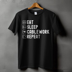 May include: Black t-shirt with the white text "EAT SLEEP CABLEWORK REPEAT" in a vertical list. The design includes icons of a burger, bed, cable plug, and a circular arrow. The shirt is hanging on a wooden hanger.