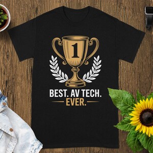 May include: Black t-shirt featuring a gold trophy graphic with the number 1 and the text "BEST. AV TECH. EVER." The trophy is surrounded by a white laurel wreath. The shirt is displayed on a wooden surface.