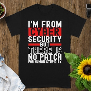 May include: Black t-shirt with the text "I'M FROM CYBER SECURITY BUT THERE IS NO PATCH FOR HUMAN STUPIDITY" in white and red lettering. The shirt is displayed on a wooden surface with a small plant and a sunflower.