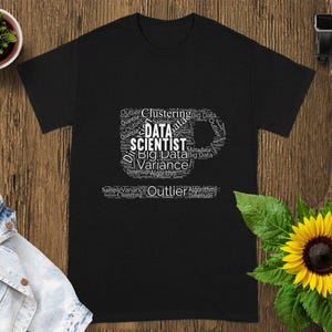 Puede incluir: Camiseta negra con un gráfico blanco en forma de taza de café, llena de términos de ciencia de datos como "Data Scientist", "Big Data" y "Variance". La camiseta se muestra sobre una superficie de madera.