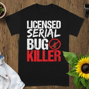 May include: Black t-shirt with the words "LICENSED SERIAL BUG KILLER" in white and red lettering. A red circle with a crossed-out bug graphic is in the center. The shirt is on a wooden surface.