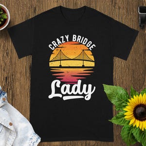 May include: A black short-sleeve t-shirt featuring a graphic of a suspension bridge silhouetted against a circular sunset gradient of yellow, orange, and red. The text "CRAZY BRIDGE Lady" is printed in white, with "CRAZY BRIDGE" arched above.