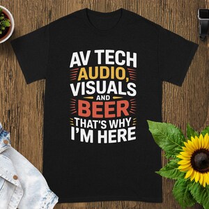 May include: Black t-shirt with the text "AV TECH AUDIO, VISUALS and BEER THAT'S WHY I'M HERE" in white, yellow, and red. The shirt is displayed on a wooden surface with a sunflower and a small potted plant.