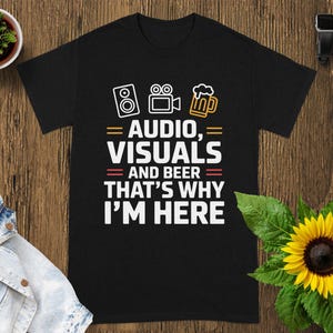 May include: Black t-shirt with white text and graphic icons related to audio, visuals, and beer. The text reads "AUDIO, VISUALS and BEER THAT'S WHY I'M HERE". The shirt is displayed on a wooden surface with a sunflower and a small plant.