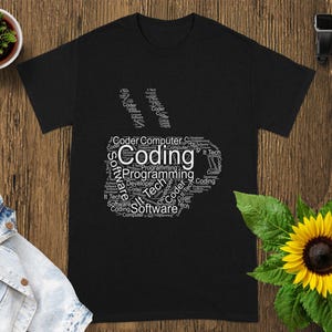 Op de afbeelding: Zwart t-shirt met een witte afbeelding in de vorm van een koffiekopje, gevuld met woorden als "Coding", "Software" en "Programming". Het shirt ligt op een houten oppervlak met een kleine potplant, een spijkerjasje, een zonnebloem en een smartwatch.