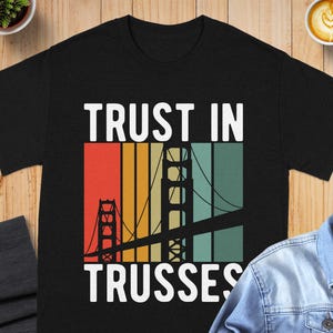 May include: A black short-sleeved t-shirt featuring a graphic print of the Golden Gate Bridge silhouette over vertical stripes in red, orange, yellow, and teal. The text "TRUST IN TRUSSES" is printed in white capital letters above and below the graphic.