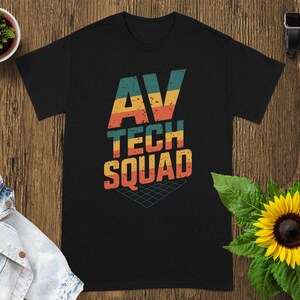 May include: Black t-shirt with the text "AV TECH SQUAD" in a retro font. The text is in orange, yellow, teal, and red. A grid graphic is below the text. A sunflower and a small potted plant are in the background.