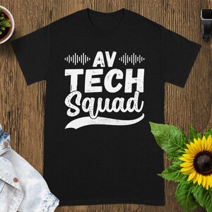 May include: Black t-shirt with the text "AV TECH Squad" in white, featuring a sound wave design above the words. The shirt is displayed on a wooden surface with a sunflower and a small plant.