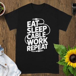 May include: Black t-shirt with the white text "EAT SLEEP CABLE WORK REPEAT". The design includes a cable graphic. The shirt is displayed on a wooden surface with a sunflower and a small potted plant.