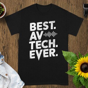May include: Black t-shirt with the text "BEST. AV TECH. EVER." in white, featuring a sound wave graphic. The shirt is displayed on a wooden surface, with a small plant and a sunflower in the background.